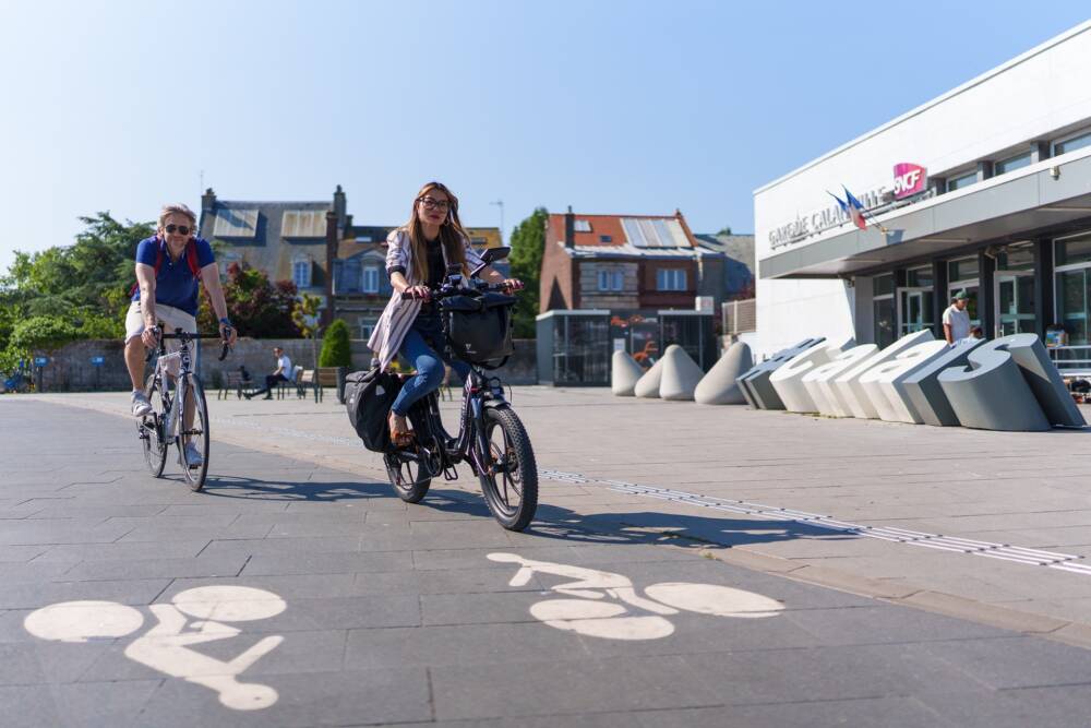 Un couple à vélo devant la gare de Calais et son espace multimodal.