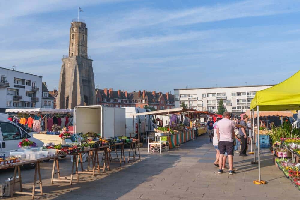 Marché animé sur la place d’Armes de Calais, dominée par la Tour du Guet, emblématique monument historique. Étals colorés de produits locaux, fruits, légumes et fleurs, attirant habitants et visiteurs dans une ambiance conviviale en plein cœur de la ville.