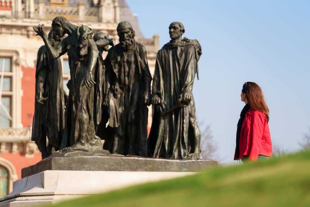 Une femme observe de près la sculpture des Bourgeois de Calais de Rodin, située devant l’Hôtel de Ville de Calais, symbole fort de l’histoire de la ville.