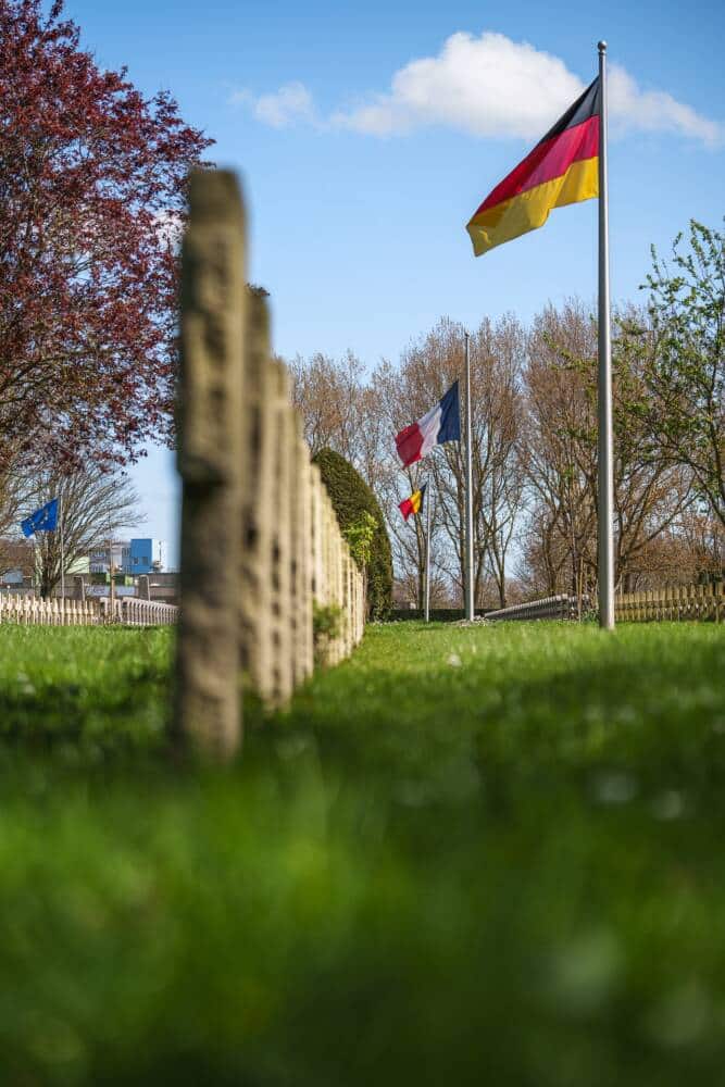 Alignement de drapeaux français, allemand, belge et européen dans les allées du cimetière Nord de Calais, symbole de mémoire et de réconciliation, sous un ciel bleu printanier.