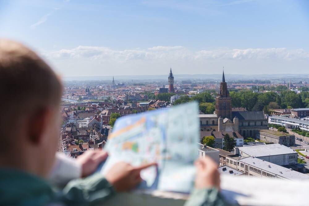 Depuis le sommet du phare de Calais, un enfant observe la ville avec une carte touristique en main, profitant d’un panorama unique sur le beffroi, l’église Notre-Dame et les toits de la ville.