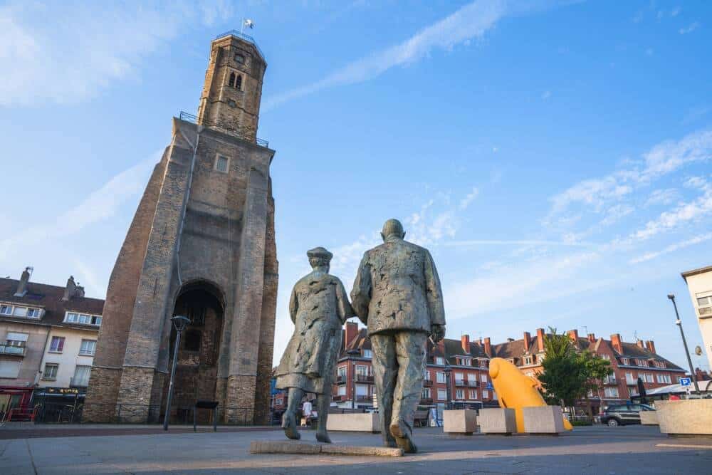 Statue de Charles de Gaulle et Yvonne Vendroux, main dans la main, au pied de la tour du Guet à Calais, sous un ciel bleu. L’œuvre symbolise l’union du couple dans la ville natale d’Yvonne, devant un monument historique emblématique du centre-ville.