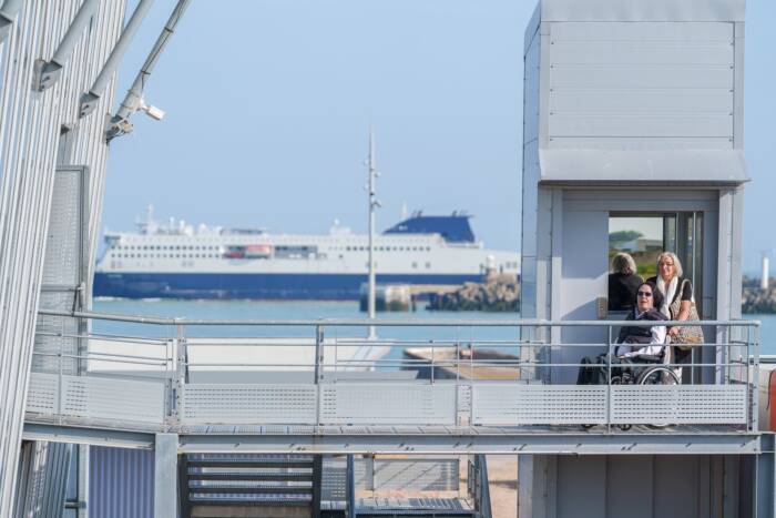 Cette photo nous montre une personne à mobilité réduite empruntant l'ascenseur pour accéder au Dragon de Calais avec vue sur un ferry en arrière plan.
