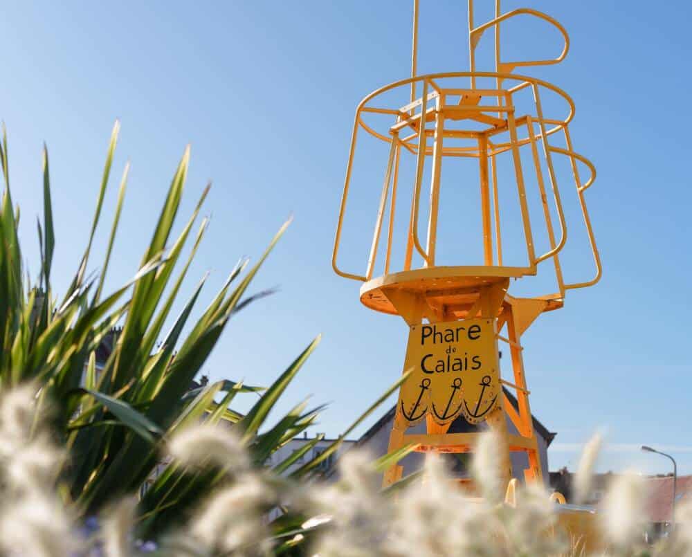 Structure métallique de signalisation jaune avec l’inscription 'Phare de Calais', installée en extérieur, entourée de végétation, sous un ciel bleu clair.