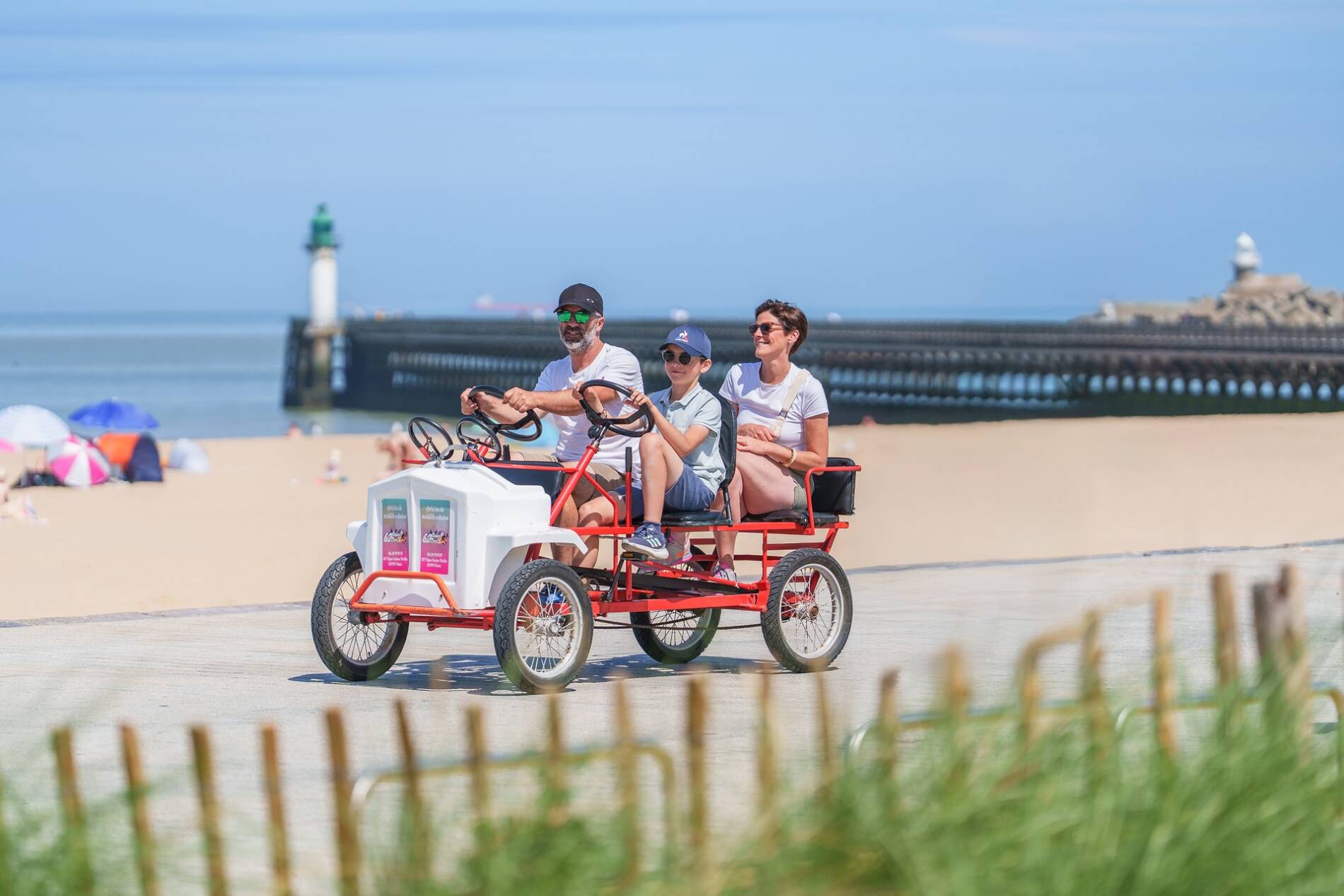 Une famille au volant d'une Rosalie sur la promenade aménagée de la plage de Calais avec la mer et la digue en arrière plan.