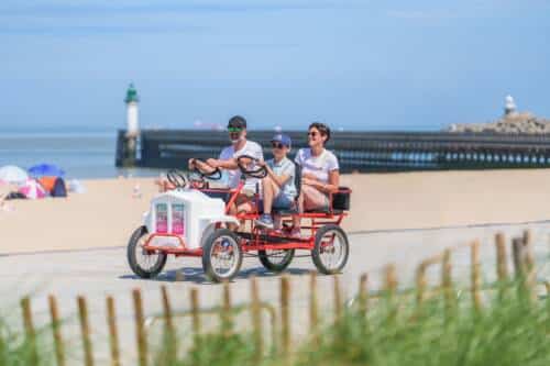 Une famille au volant d'une Rosalie sur la promenade aménagée de la plage de Calais avec la mer et la digue en arrière plan.