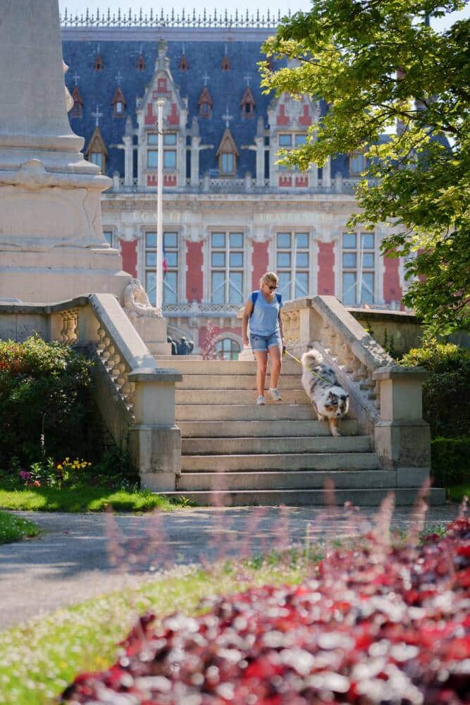 Une femme promenant son chien dans le Parc Sain-Pierre avec en arrière-plan, l'hôtel de ville de Calais.