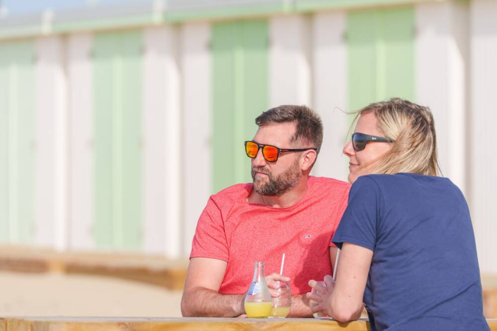 Un couple attablé sur la plage des baraques avec ses chalets en arrière plan.